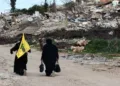 Women walk near destroyed buildings, with one holding the flag of Hezbollah, in the southern Lebanese village of Kfar Kila