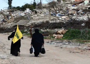 Women walk near destroyed buildings, with one holding the flag of Hezbollah, in the southern Lebanese village of Kfar Kila