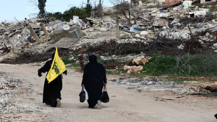 Women walk near destroyed buildings, with one holding the flag of Hezbollah, in the southern Lebanese village of Kfar Kila